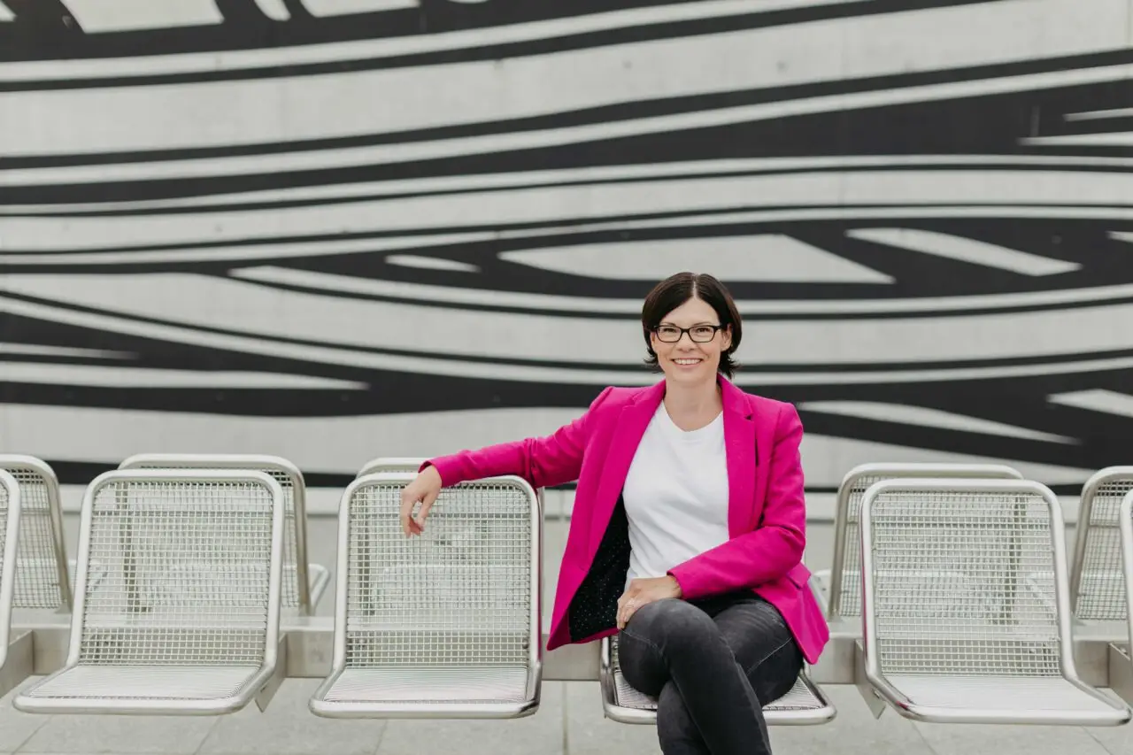 Professionelle Businessfrau mit pinkem Blazer sitzt entspannt auf Metallstühlen vor moderner Architektur.