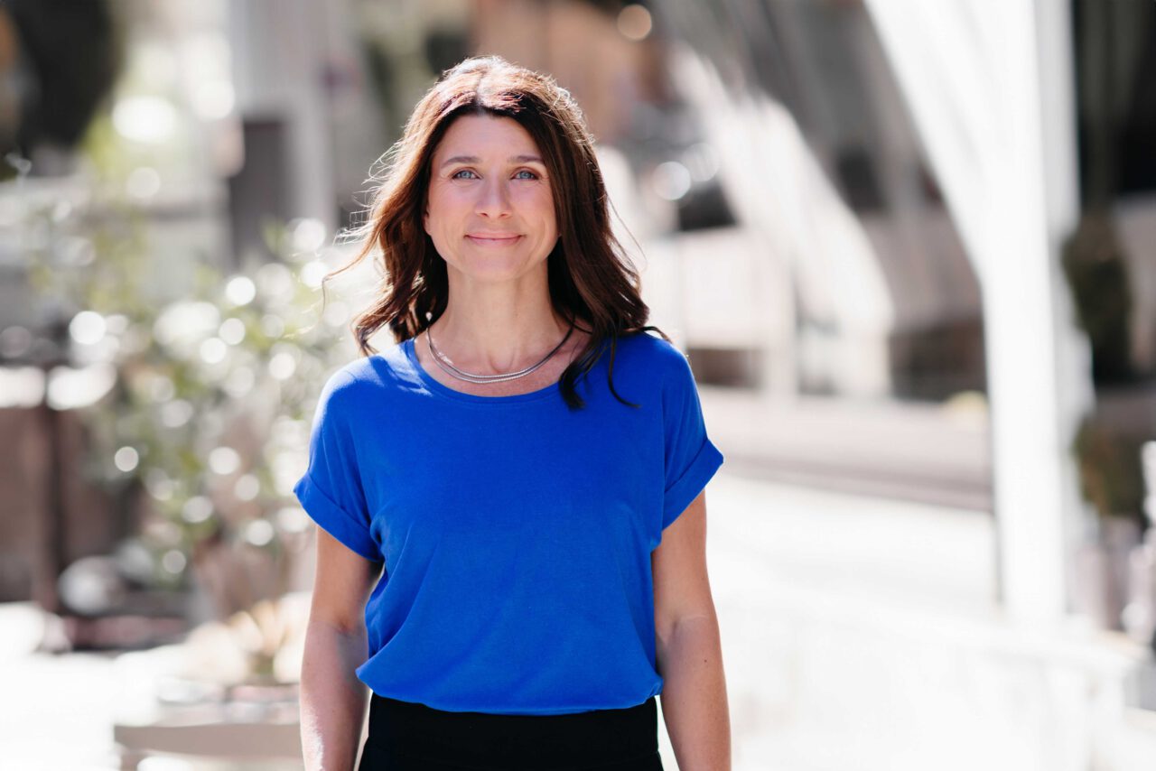 Lächelnde Frau in blauem Shirt mit langen braunen Haaren, steht entspannt im Sonnenlicht auf einer urbanen Straße.