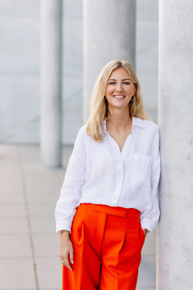 Junge Unternehmerin in weißer Bluse und roter Hose lehnt lächelnd an einer Betonsäule – modernes Personal Branding Portrait von RAUMFOTOGRAFIN Barbara Meyer-Selinger aus München.