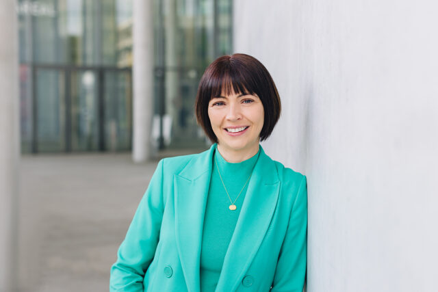 Frau mit kurzem dunklem Bob trägt einen grünen Blazer über einem grünen Oberteil und lehnt lächelnd an einer hellen Wand vor einer modernen Glasfassade.