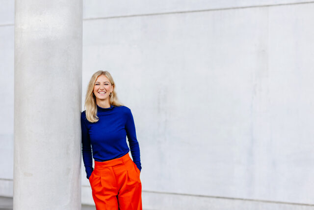 Blonde Frau in blauem Oberteil und orangefarbener Hose lehnt lächelnd an einer hellen Betonsäule vor moderner Architektur.