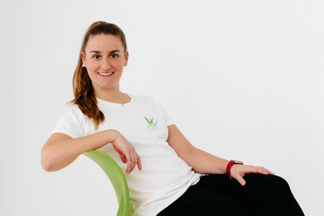 rau mit zusammengebundenen braunen Haaren sitzt seitlich auf einem grünen Stuhl, trägt ein weißes T-Shirt und eine schwarze Hose und lächelt vor weißem Hintergrund in die Kamera.