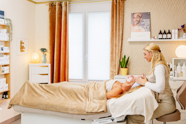 Kosmetikerin mit zusammengebundenen blonden Haaren trägt eine Kundin mit aufgetragener Gesichtsmaske auf einer Behandlungsliege in einem warm beleuchteten Kosmetikstudio mit Vorhängen, Pflegeprodukten und dekorativen Elementen.