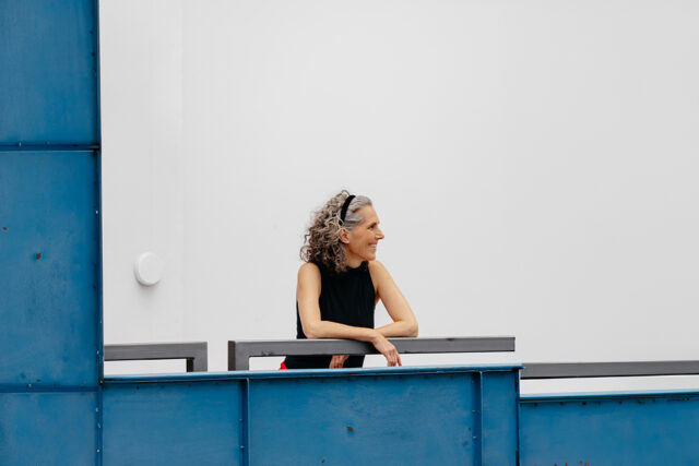 Frau mit grauen lockigen Haaren und schwarzem ärmellosem Oberteil lehnt seitlich an einem blauen Geländer und blickt lächelnd nach rechts vor einer weißen Wand.