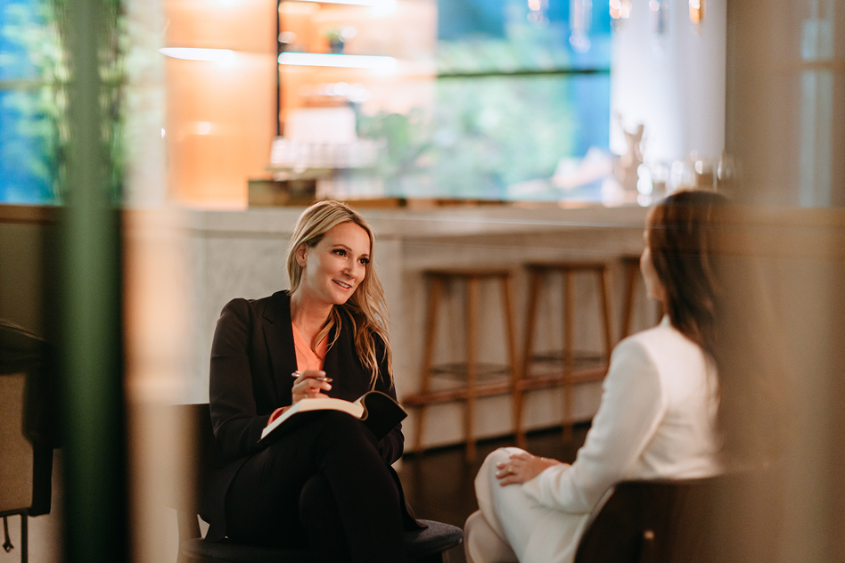 Zwei Frauen sitzen sich in einer modernen Lounge gegenüber, eine blonde Frau im dunklen Blazer hält ein Notizbuch und spricht lächelnd mit einer Frau in hellem Anzug.
