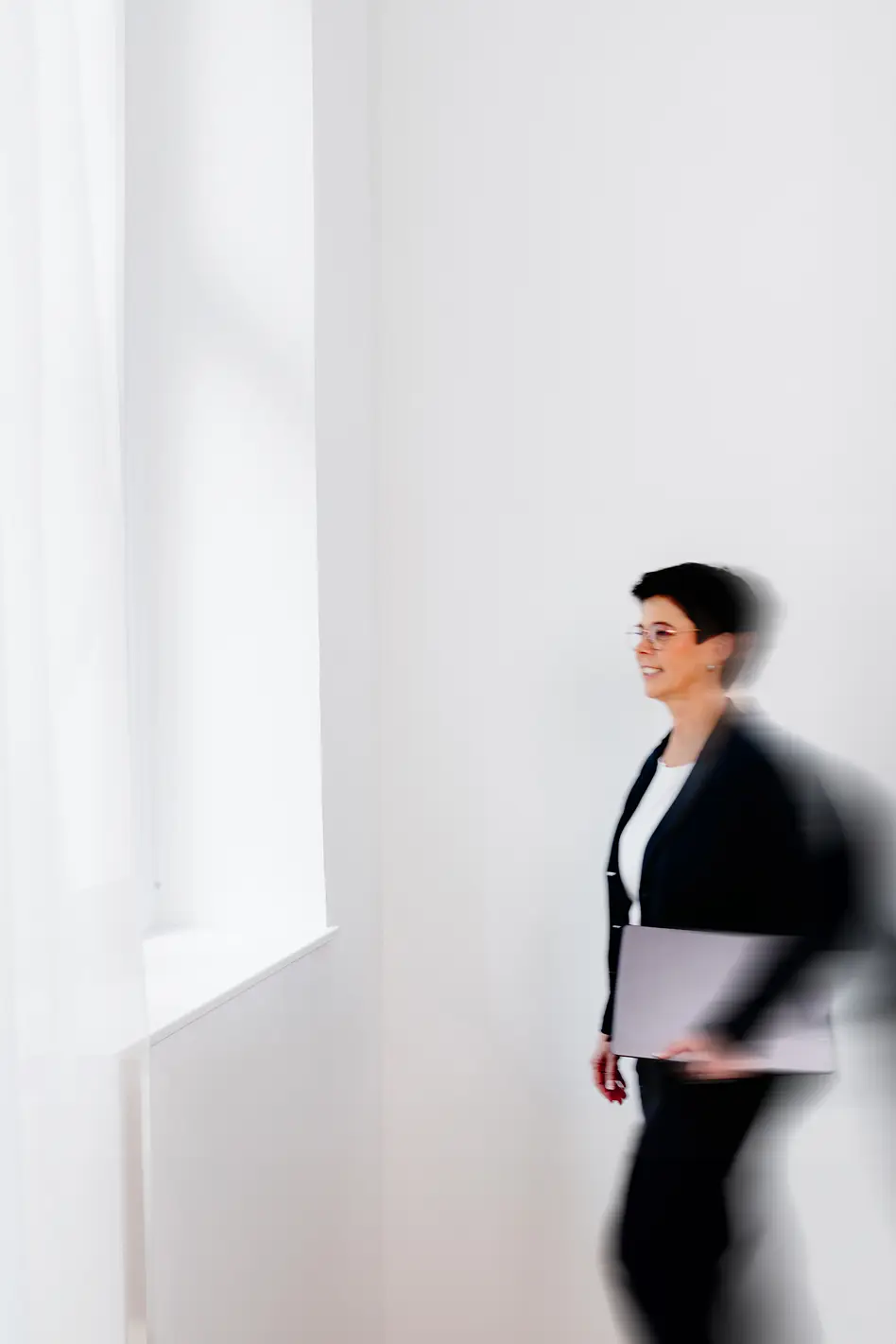 Frau mit kurzen dunklen Haaren, Brille und schwarzem Blazer läuft mit Laptop unter dem Arm durch einen hellen Raum mit großen Fenstern.