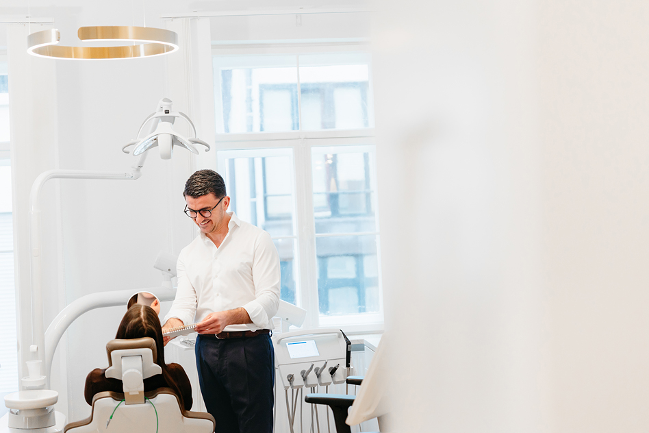 Zahnarzt mit Brille und weißem Hemd steht neben einer Patientin, die in einem Behandlungsstuhl sitzt, und erklärt ihr anhand eines Modells oder einer Karte eine Behandlung in einer hellen modernen Zahnarztpraxis mit großen Fenstern und Behandlungsleuchte.